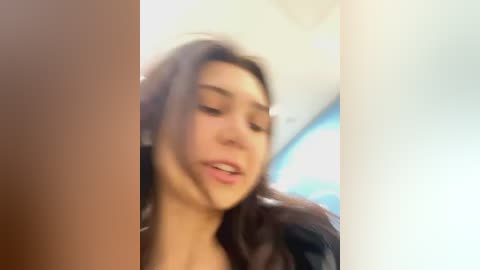 Media: Blurry video of a young woman with long brown hair and light skin, wearing a black jacket, captured from a slightly low angle. The background is indistinct, possibly an indoor setting with soft lighting.