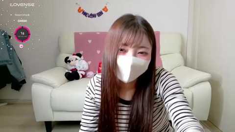 Media: A video of an Asian woman with long brown hair wearing a face mask, black and white striped shirt, and sitting on a white leather couch in a pink room with a plush toy panda.