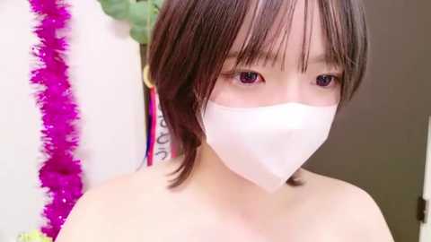 Media: Video of an Asian woman with short, dark hair, wearing a white face mask, standing against a backdrop of pink streamers and a white wall.