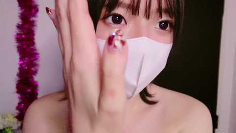 Media: Video of an Asian woman with light skin, wearing a white mask, touching her nose with a long fingernail, in a room with purple tinsel and a white door in the background.