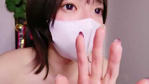 Media: Video of a young Asian woman with straight black hair, wearing a white mask, with a hand gesturing from the right side, in a minimalist room.