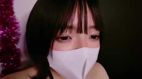 Media: Video of an Asian woman with dark hair, wearing a white mask, looking downcast. Background is dark and blurry, with a hint of a red tinsel decoration on the left.