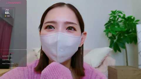 Media: Video of an Asian woman with straight, shoulder-length brown hair, wearing a white face mask and a pink sweater. She has a calm expression and is indoors, with a green plant in the background.