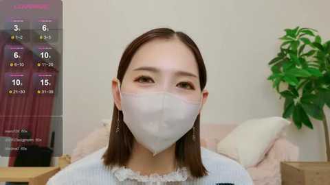 Media: Video of an East Asian woman with shoulder-length brown hair, wearing a white face mask, a light blue sweater, and gold earrings, sitting indoors with a green plant and a pink curtain in the background.