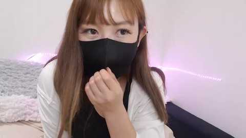 Media: Video of an Asian woman with long brown hair, wearing a black face mask, white top, and a black vest, covering her mouth with her right hand. Background features a plush, white rug and a white wall with soft purple lighting.