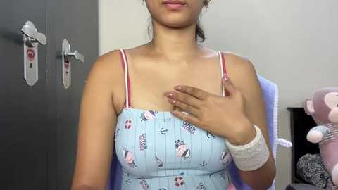 Media: A video of a young woman with light brown skin, wearing a light blue, cartoon-print dress with thin straps, touching her chest. She has a slender build and her hair is tied back. The background features a gray wardrobe and plush toys.