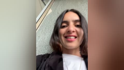 Media: Video of a young woman with long dark hair, wearing a black jacket over a white shirt, smiling broadly against a textured light green wall.