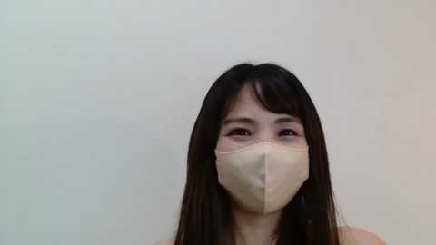 Media: Video of an Asian woman with long black hair, wearing a beige surgical mask, against a plain light beige background.