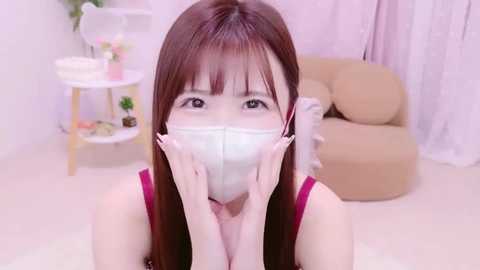 Media: Video of an Asian woman with long brown hair, wearing a face mask, red tank top, and white gloves, in a pastel-colored room with a beige couch and white furniture.