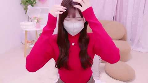 Media: Video of an Asian woman with long brown hair, wearing a red sweater, a white mask, and a heart pendant, adjusting her hair in a pastel-pink room with beige cushions and a potted plant.