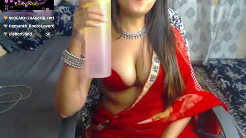 Media: Video of a South Asian woman with long black hair, wearing a red sari with a low-cut blouse, drinking from a glass, surrounded by colorful cushions and a patterned curtain.