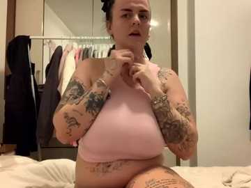 Media: A video of a tattooed woman with dark hair, wearing a pink sports bra, adjusting her bra straps in a bedroom with clothes hanging in a closet.