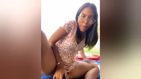 Media: A video of a young, slender woman with medium-length black hair, wearing glasses and a floral top, sitting on a blue towel outdoors, partially obscured by a blurry figure.