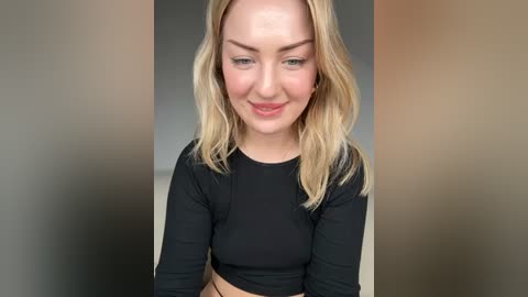 Media: Video of a blonde Caucasian woman with fair skin, wearing a black long-sleeve crop top, smiling gently. Background is a blurred gradient of beige, brown, and gray.