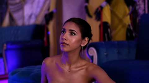 Media: Video of a topless, slender woman with olive skin, dark hair in an updo, wearing dangling earrings, standing in a dimly lit room with blue sofas and colorful jackets hanging on the wall.