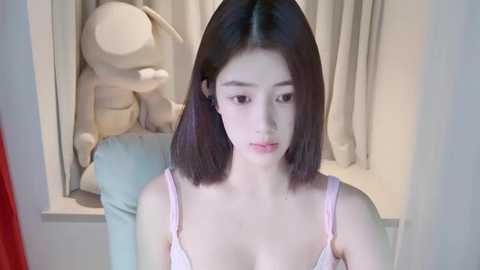 Media: A video of a young Asian woman with straight black hair, fair skin, and a slender physique, wearing a light pink spaghetti-strap top, standing in front of a white window with light curtains.