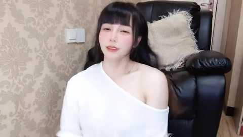 Media: Video of a young Asian woman with long black hair, fair skin, and a slim build, wearing a white off-shoulder top, sitting in a black leather armchair with beige floral wallpaper in the background.