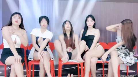 Media: Video of five East Asian women, diverse in body types, wearing revealing black and white dresses, seated on red chairs in a modern indoor setting with white curtains and spotlights.