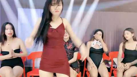 Media: Video of five Asian women in black lingerie on red chairs, with one woman in a red dress center foreground, in a brightly lit, modern indoor setting.