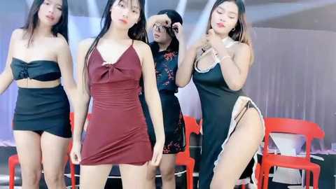 Media: Video of four Asian women in provocative black dresses and high heels, posing confidently in a modern, minimalist room with red chairs and white curtains.