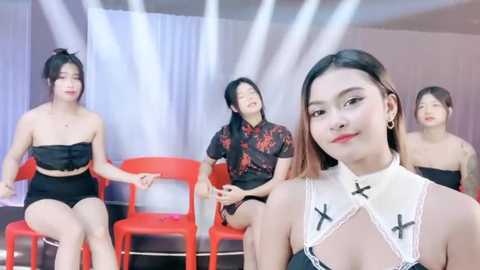 Media: Video of five Asian women, including a center woman with shoulder-length brown hair, wearing a white choker with black crosses, sitting on red chairs in a studio.