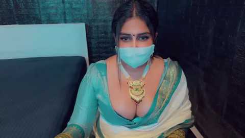 Media: Video of a South Asian woman with fair skin, dark hair, wearing a green and white saree, gold necklace, and a face mask, sitting on a bed with dark, patterned walls in the background.