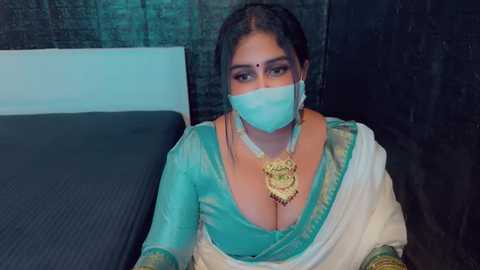 Media: Video of a South Asian woman in a teal and white saree, with a gold necklace, wearing a face mask, sitting on a black cushioned bed against a dark backdrop.