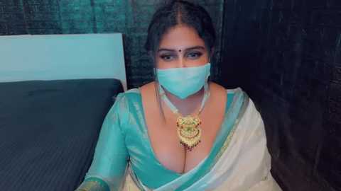 Media: A video of a woman with dark skin and dark hair, wearing a turquoise and white saree, a gold necklace, and a face mask, seated indoors against a dark backdrop.