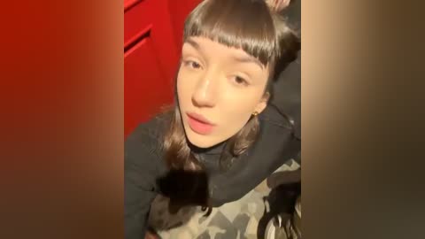 Media: Video of a young woman with straight, blunt bangs, light brown skin, and wearing a black turtleneck sweater. She has a neutral expression, standing in front of a red door and beige walls.