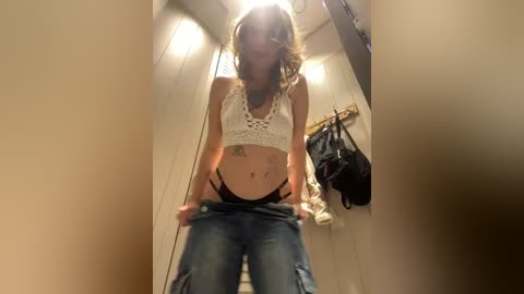 Media: Video of a young woman with light skin, wearing a white crochet crop top and blue jeans, looking down at her crotch. Background includes a beige wall and a black handbag hanging on a hook.