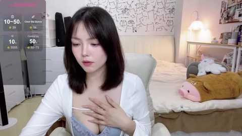 Media: Video of a young Asian woman with shoulder-length black hair, wearing a white cardigan and a revealing blue lace bra, seated in a cozy, cluttered bedroom with a white bed, stuffed animals, and a digital clock.