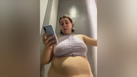 Media: A candid video of a plus-sized woman with fair skin, wearing a light pink plaid crop top and no pants, taking a selfie in a mirror-walled changing room.