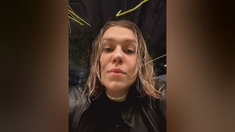 Media: Video of a young woman with wet, shoulder-length blonde hair, wearing a black leather jacket, standing in a dark, possibly urban setting.