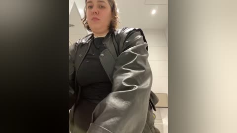 Media: Video of a plus-sized woman with curly brown hair, wearing a black leather jacket and a black top, standing in a modern, white-walled room.