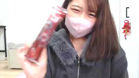 Media: Video of a young Asian woman with straight brown hair, wearing a pink surgical mask and a dark coat with a gray fur collar. She holds a red, checkered candy. Background includes a white wall with a red Christmas decoration and a piece of furniture.