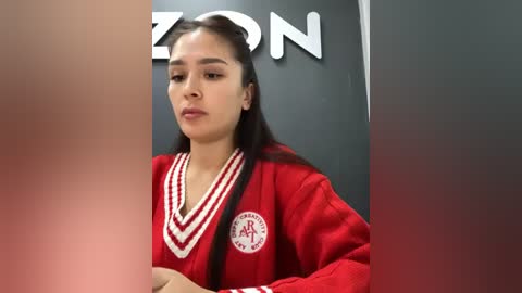 Media: Video of a young woman with straight, dark hair, wearing a red sweater with white stripes and a school logo, standing in a room with a gray wall and partially visible \"Amazon\" signage.