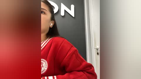 Media: Video of a young woman with long, dark hair in a red, striped, long-sleeved sweater, partially framed by a door. Background shows a black wall with white letters partially visible.