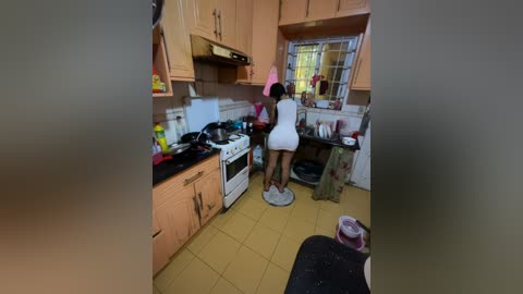 Media: A video captures a woman in a white mini-dress and pink headband cooking in a cramped, cluttered kitchen with wooden cabinets and yellow tile floors.