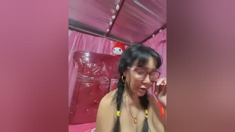 Media: Video of a young Asian woman with black hair in pigtails, wearing glasses and a black bikini, sitting in a pink room with a red inflatable bed and a small plush toy.