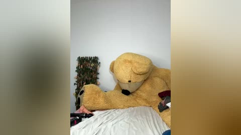 Media: Video of a large, beige teddy bear lying on a messy bed with white sheets, surrounded by a green artificial plant and a blurred person.