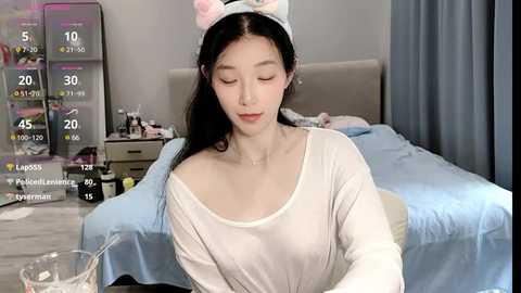 Media: Video of a young East Asian woman with long black hair and a white bunny headband, wearing a white off-shoulder top, sitting in a bedroom with a blue bed, a nightstand, and a temperature display.