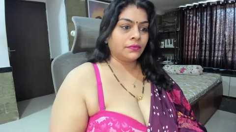 Media: Video of a plus-size South Asian woman with long black hair, wearing a bright pink patterned sari, sitting on a beige chair in a modern bedroom with a bed and dark curtains.