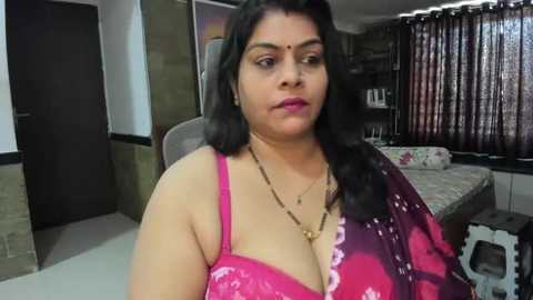 Media: Video of a middle-aged South Asian woman with medium skin tone, wearing a bright pink saree, black bindi, and gold necklace. She is seated in a modestly furnished bedroom with a bed, curtains, and wall art in the background.