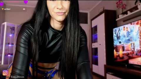 Media: A video of a young woman with long black hair, wearing a black leather crop top, standing indoors near a TV showing a movie.