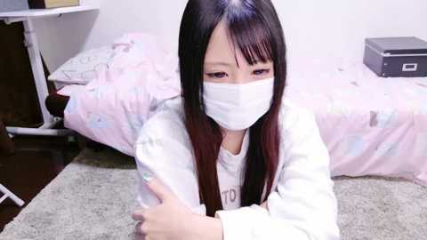 Media: Video of an Asian woman with long black hair, wearing a white mask, lying on a grey carpet in a simple bedroom with a pink bedspread and a black box.