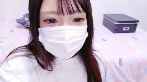 Media: Video of an East Asian woman with long, straight dark hair, wearing a white face mask, sitting on a pink bed with polka dot sheets, against a plain white background.