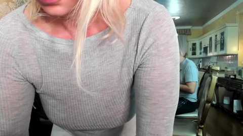 Media: Video of a blonde woman with fair skin wearing a tight, ribbed gray top, leaning over a kitchen counter. Behind her, a man with short hair and a light blue shirt sits at a wooden table.