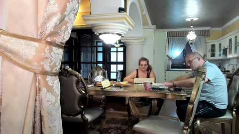 Media: Video of a man and woman in a lavish, ornate dining room, wearing casual clothes, with a wooden table set for a meal, surrounded by intricate decor.