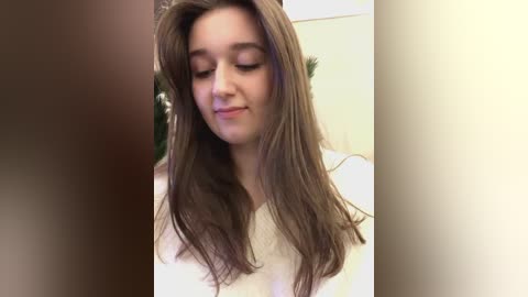 Media: Video of a young woman with fair skin, long brown hair, and closed eyes, wearing a white top. Background features blurred greenery.