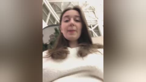 Media: Video of a young woman with long brown hair, wearing a white sweater, standing indoors with a blurred background featuring a staircase and potted plant.